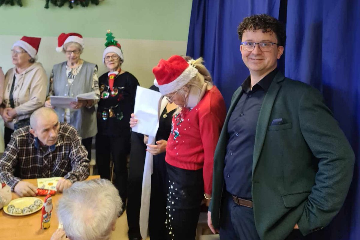 Kilka osób w czapkach Mikołaja stoi obok stołu z jedzeniem, w centrum mężczyzna w ciemnej marynarce.