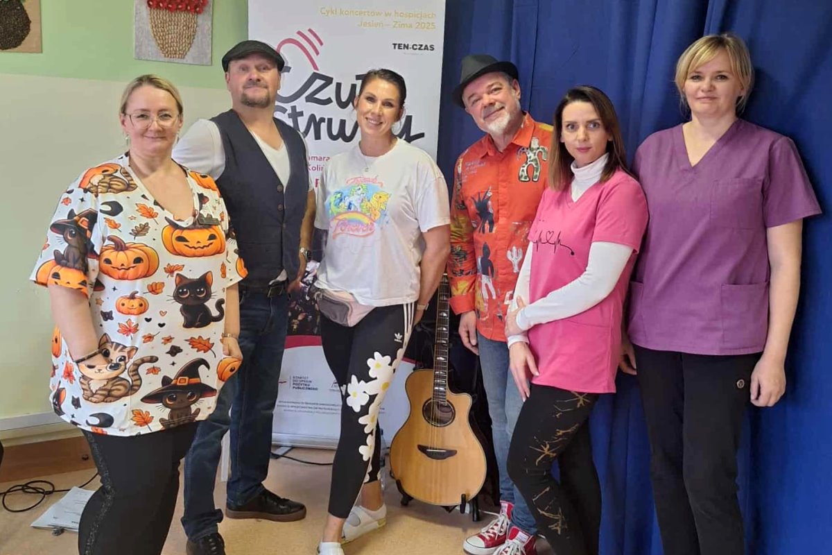 Grupa os&oacute;b pozuje do zdjęcia na tle niebieskiej zasłony. Na pierwszym planie widać gitarę, a wszyscy uśmiechają się do aparatu.