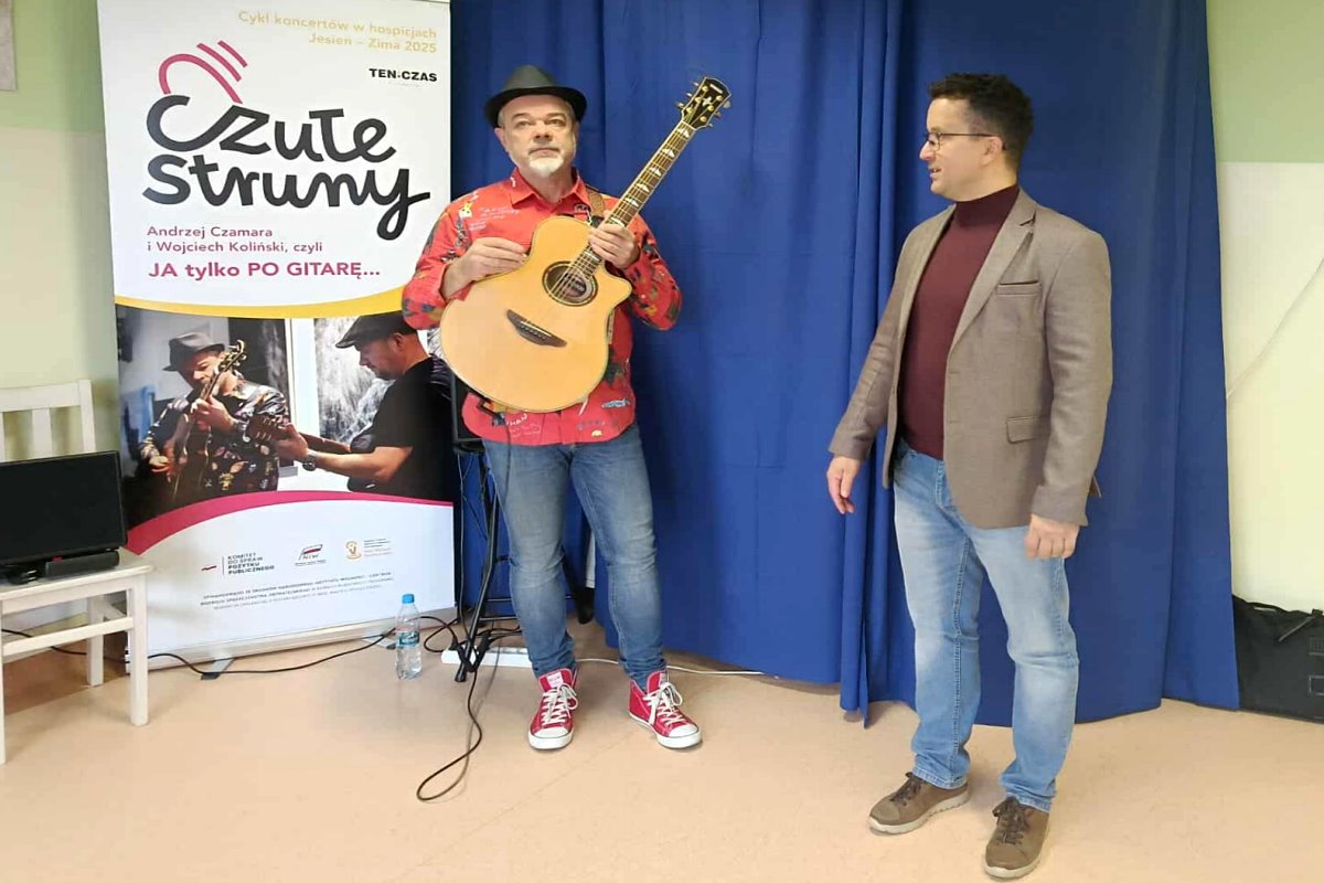Mężczyzna z gitarą stoi obok drugiego mężczyzny. Za nimi znajduje się baner z napisem &bdquo;Czułe Struny&rdquo;.