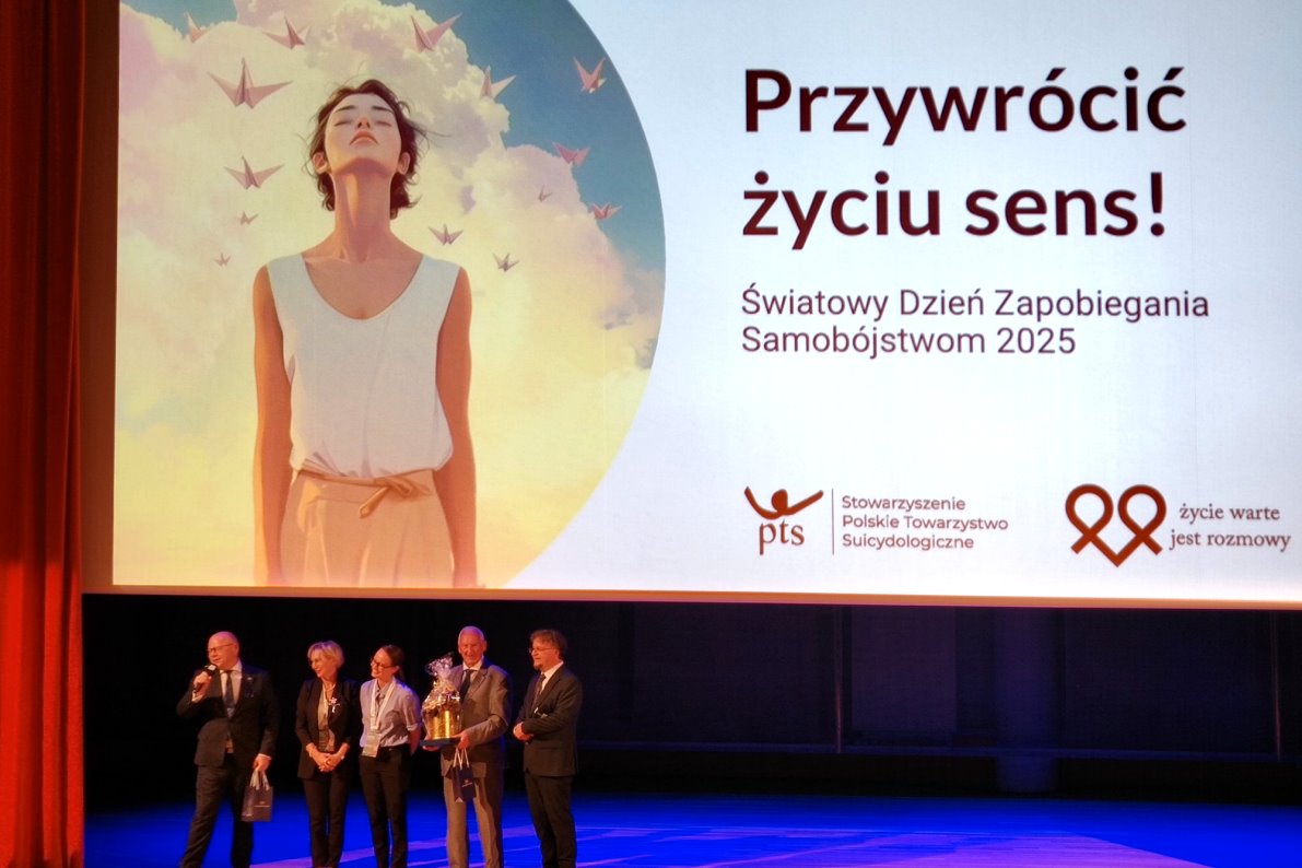 Na scenie stoi kilka os&oacute;b przed dużym ekranem z napisem &bdquo;Przywr&oacute;cić życiu sens! Światowy Dzień Zapobiegania Samob&oacute;jstwom 2025&rdquo;. Na ekranie widoczna jest grafika kobiety w świetle słońca.