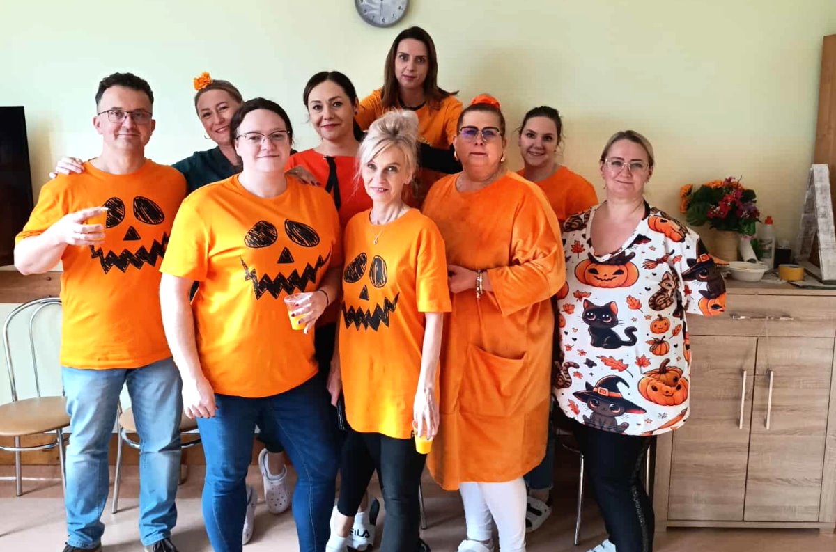 Grupa osób stoi razem w pokoju. Część z nich ma pomarańczowe koszulki z namalowanymi dyniami, jak na Halloween. Wszyscy się uśmiechają.
