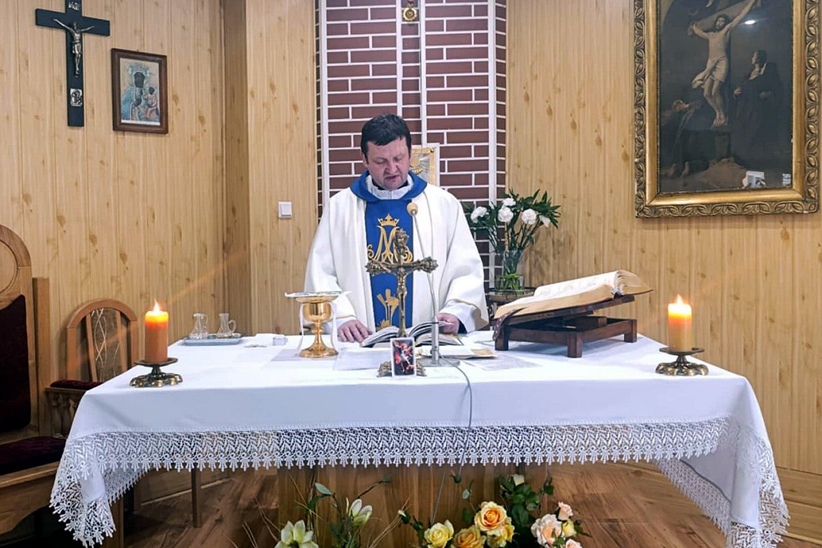 Kapłan w szatach liturgicznych stoi przy ołtarzu podczas nabożeństwa, na stole świece i księga, wnętrze drewnianej kaplicy.